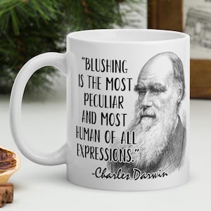 Può includere: Una tazza in ceramica bianca con un'illustrazione in bianco e nero di Charles Darwin e una sua citazione che recita: "Arrossire è la cosa più peculiare e più umana di tutte le espressioni." -Charles Darwin