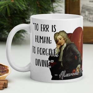 May include: White ceramic coffee mug with a black quote that reads "To err is human; to forgive, divine." - Alexander Pope. The mug features a portrait of Alexander Pope in a red chair.