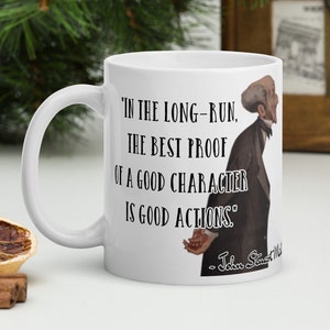 May include: White ceramic coffee mug with a black quote that reads "In the long-run, the best proof of a good character is good actions." The quote is attributed to John Stuart Mill. A black and white illustration of a man is on the right side of the mug.