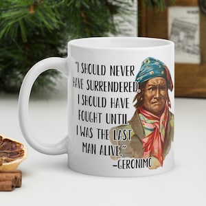 Op de afbeelding: Witte keramische koffiemok met een zwart-wit citaat van Geronimo: "Ik had me nooit moeten overgeven. Ik had moeten vechten tot ik de laatste man in leven was." Het citaat wordt omringd door een vintage illustratie van Geronimo.