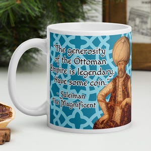 Peut inclure: Une tasse à café en céramique blanche avec un motif bleu et blanc. La tasse présente une citation de Soliman le Magnifique : « La générosité de l'Empire ottoman est légendaire. Prenez un peu de monnaie. »