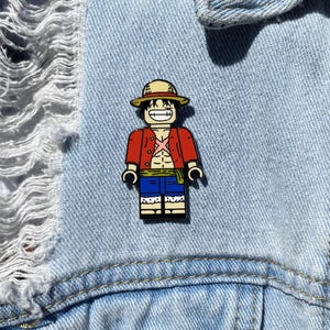 Peut inclure: Une épingle en émail colorée représentant un personnage de dessin animé en veste rouge, pantalon bleu et chapeau de paille. L'épingle est conçue dans un style de blocs, rappelant les blocs de construction, sur un fond en jean bleu clair.