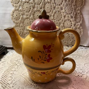 May include: A yellow ceramic teapot and teacup set. The teapot has a burgundy lid with white polka dots and a green finial. Red floral designs and white dots decorate the teapot and teacup. The set is on a white lace tablecloth.