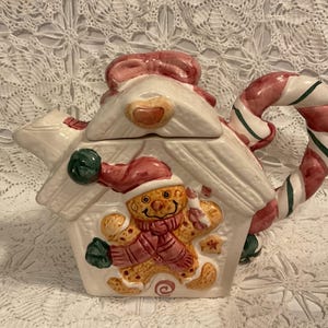 May include: A ceramic Christmas teapot shaped like a gingerbread house. The white teapot features a gingerbread man, a candy cane handle, and a pink bow on top. The gingerbread man wears a red hat and scarf.