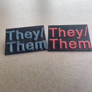 May include: Two rectangular badges with the text "They/ Them". One badge has blue text on a black background, and the other has red text on a black background. The badges are likely for expressing pronouns.