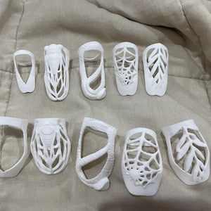 May include: A collection of ten white, 3D-printed nail covers featuring intricate cut-out designs. The set includes patterns such as a spider web, a snake, a cicada, and leaf-like motifs, each shaped to fit over a fingernail with an open-frame structure.