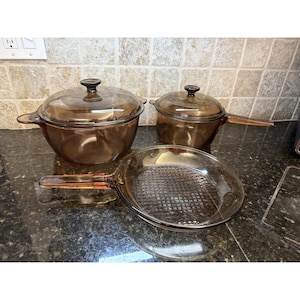 May include: A set of three amber-colored glass cookware pieces including a large round pot with a lid, a smaller saucepan with a lid, and a shallow frying pan. Each piece is made of transparent brown glass with integrated handles and lids.