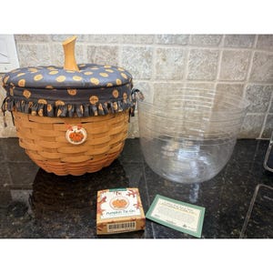 May include: A pumpkin-shaped basket with a wooden stem and a fabric lid featuring gold polka dots. A clear plastic bowl and a small box labeled "Pumpkin Tie-On" are also present. The basket has a pumpkin-themed tag.