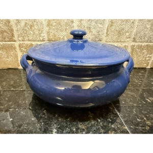May include: A blue ceramic casserole dish with a lid. The dish has two handles and a knob on the lid. The dish is a solid blue color and appears to be glazed. The dish is sitting on a dark surface.