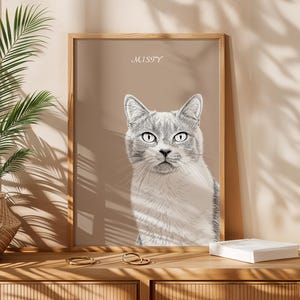 May include: A framed portrait of a gray and white cat with the name "MISTY" above it. The artwork is displayed on a wooden surface with a woven basket and a plant in the background.