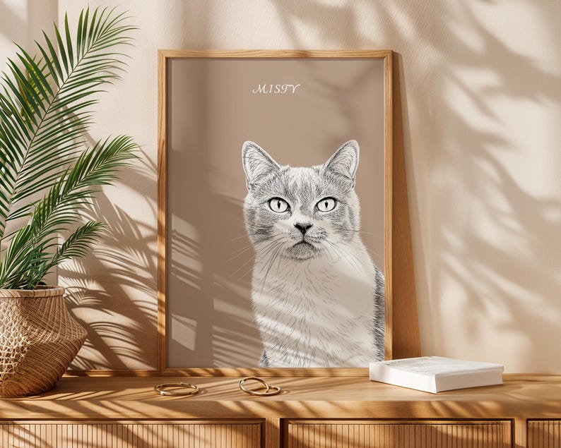 May include: A framed portrait of a gray and white cat with the name "MISTY" above it. The artwork is displayed on a wooden surface with a woven basket and a plant in the background.