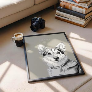 May include: A framed black and white portrait of a dog with the name "Maple" above the image. The dog has a collar and is smiling. A black camera, a coffee mug, and a stack of books are also in the image.