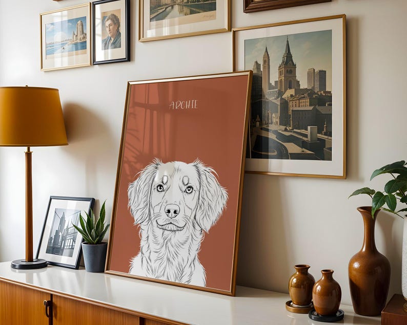 May include: A framed art print featuring a detailed black and white illustration of a dog's face, with the name "Archie" above. The print has a rust-colored background and a gold frame. Other framed art and decor are visible.