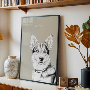 May include: Framed black and white dog portrait with the name "MAPLE" in a script font. The dog has blue eyes and a collar. The artwork is displayed on a white surface with a lamp, vase, and decorative items.