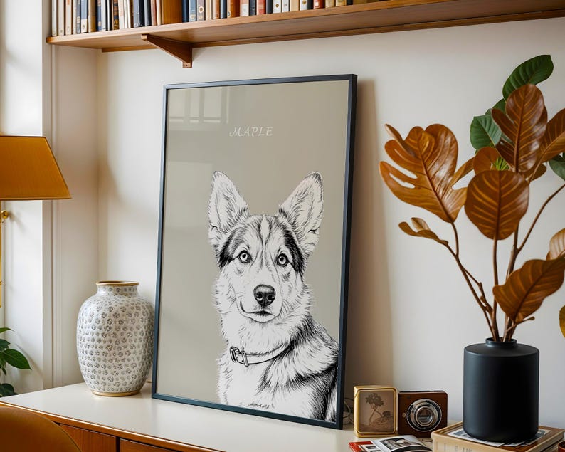 May include: Framed black and white dog portrait with the name "MAPLE" in a script font. The dog has blue eyes and a collar. The artwork is displayed on a white surface with a lamp, vase, and decorative items.