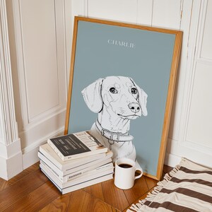 May include: A framed art print featuring a sketched portrait of a dachshund dog named "Charlie" on a muted blue background. The artwork is in a light wood frame and is propped against a wall next to a stack of books and a white mug.
