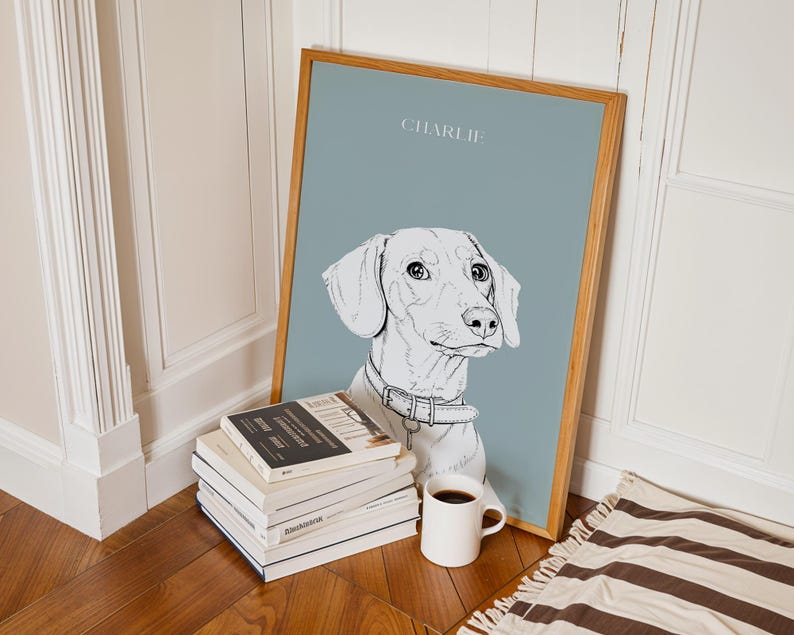 May include: A framed art print featuring a sketched portrait of a dachshund dog named "Charlie" on a muted blue background. The artwork is in a light wood frame and is propped against a wall next to a stack of books and a white mug.