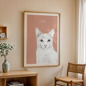 May include: A framed art print featuring a detailed pencil sketch of a cat's face, set against a muted rose background. The cat's name, "LOUIE," is printed at the top. The artwork is displayed in a room with a wooden chair and a side table.
