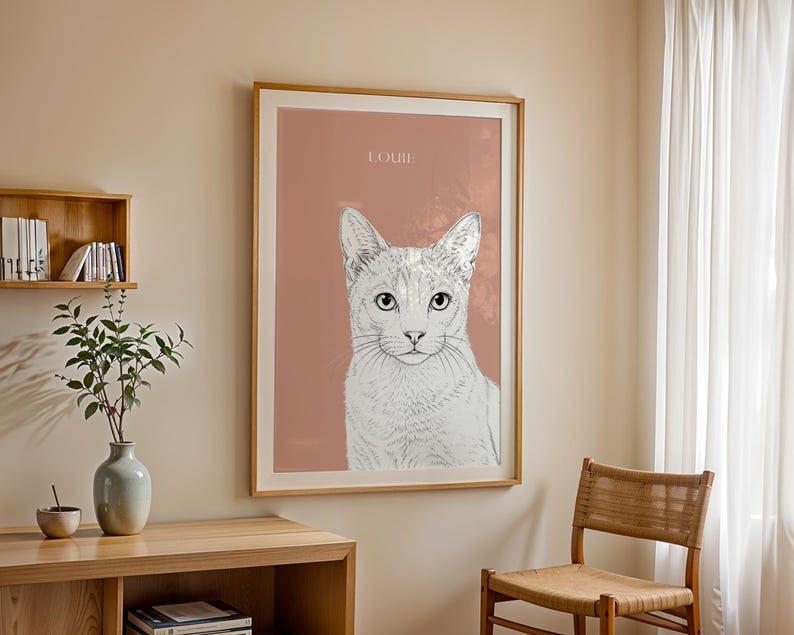 May include: A framed art print featuring a detailed pencil sketch of a cat's face, set against a muted rose background. The cat's name, "LOUIE," is printed at the top. The artwork is displayed in a room with a wooden chair and a side table.