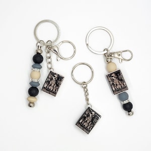 May include: Three keychains with silver-tone rings and clasps. Each keychain features a small, rectangular charm with the text "THE RITUAL" and a skull image. Two keychains have beaded accents in black, gray, and cream colors.