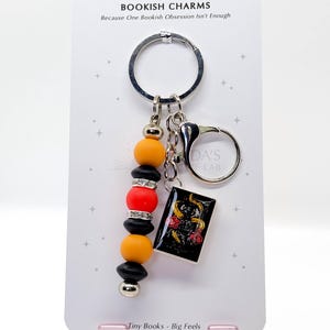 May include: A keychain with a black, silver, orange, and red beaded charm and a small book charm. The book charm has a black cover with a floral design. The keychain also has the text "BOOKISH CHARMS" and "Tiny Books - Big Feels".