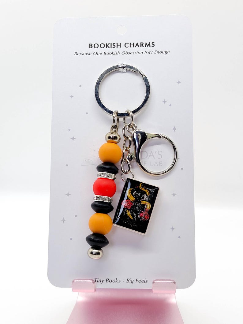 May include: A keychain with a black, silver, orange, and red beaded charm and a small book charm. The book charm has a black cover with a floral design. The keychain also has the text "BOOKISH CHARMS" and "Tiny Books - Big Feels".
