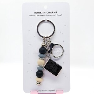 May include: A keychain with a beaded charm and a small book charm. The charm features black, gray, and cream-colored beads, with a silver accent. The text on the card reads "BOOKISH CHARMS" and "Tiny Books - Big Feels."