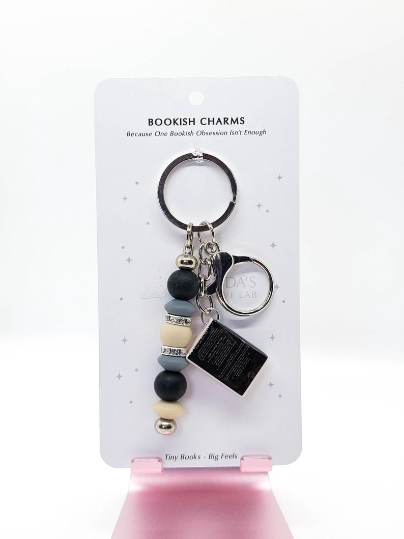 May include: A keychain with a beaded charm and a small book charm. The charm features black, gray, and cream-colored beads, with a silver accent. The text on the card reads "BOOKISH CHARMS" and "Tiny Books - Big Feels."