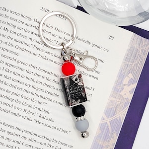 May include: A silver keychain with a charm featuring a miniature book, black and gray beads, and a red bead. The book charm reads "Haunting Adeline" and is attached to a silver clasp and keyring. The keychain rests on an open book.