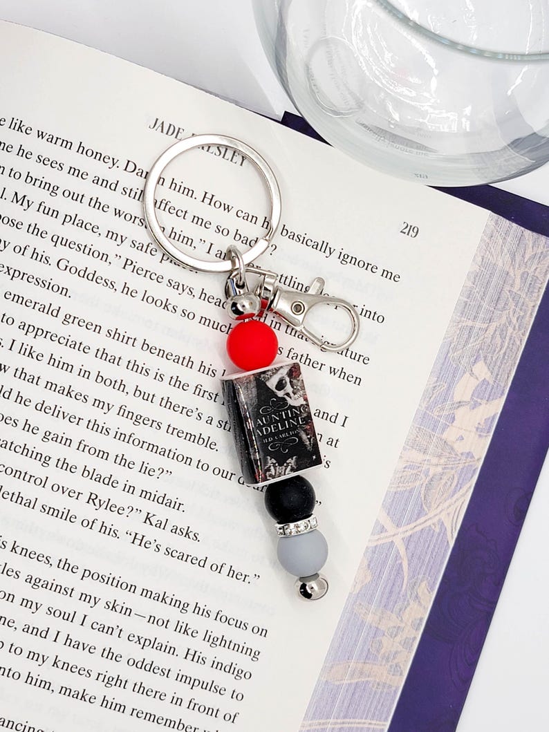 May include: A silver keychain with a charm featuring a miniature book, black and gray beads, and a red bead. The book charm reads "Haunting Adeline" and is attached to a silver clasp and keyring. The keychain rests on an open book.