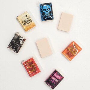 May include: Miniature book charms with titles like "Fourth Wing," "Lights Out," and "Court of Thorns and Roses." Each charm features a book cover design and a silver-colored keyring. The charms are in various colors, including red, orange, and black.