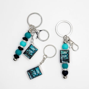 May include: Three keychains with silver rings and clasps. Each keychain features a charm with the text "LEATHER & LARK" in teal and black. The keychains also have teal, black, and silver beads.