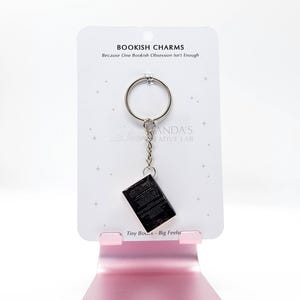 May include: A keychain featuring a miniature black book charm. The charm hangs from a silver ring and chain, attached to a white card with the text "BOOKISH CHARMS" and "Because One Bookish Obsession Isn't Enough". The card is displayed on a pink stand.