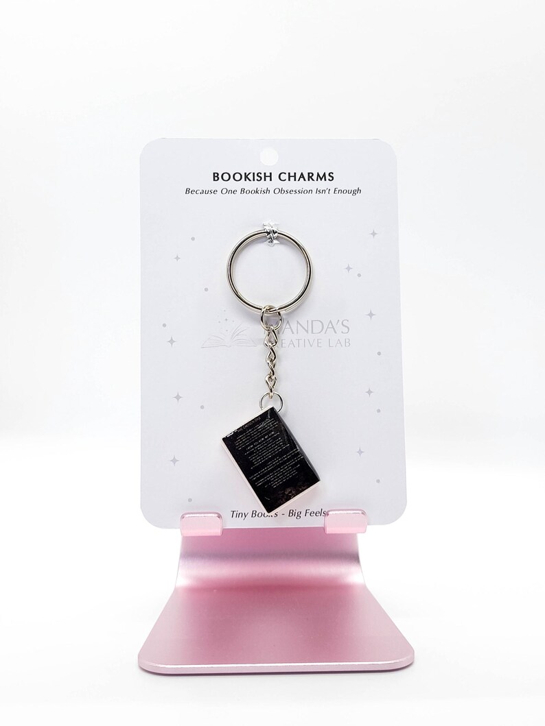 May include: A keychain featuring a miniature black book charm. The charm hangs from a silver ring and chain, attached to a white card with the text "BOOKISH CHARMS" and "Because One Bookish Obsession Isn't Enough". The card is displayed on a pink stand.
