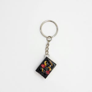 May include: Black rectangular keychain with a silver chain and ring. The keychain features a design of a yellow snake, red roses, and gold accents on a black background. The keychain is a decorative accessory.