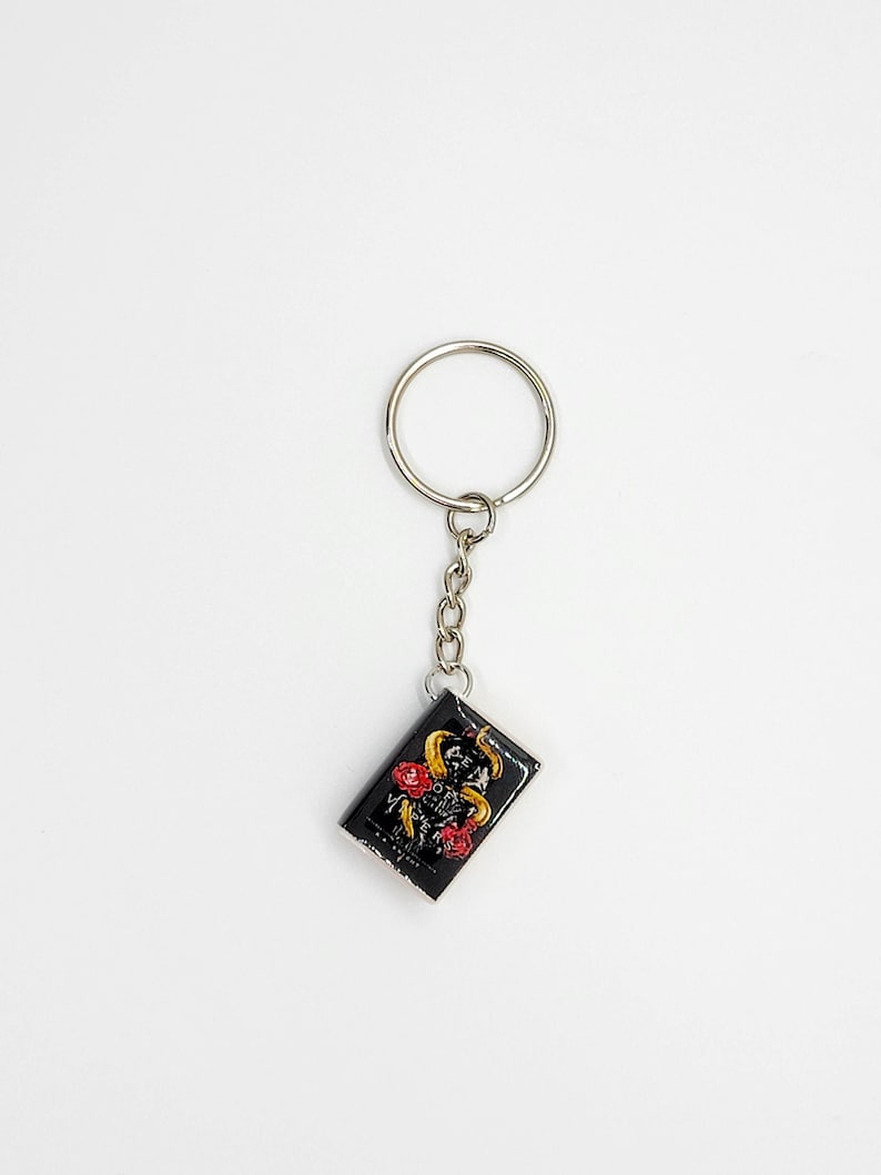 May include: Black rectangular keychain with a silver chain and ring. The keychain features a design of a yellow snake, red roses, and gold accents on a black background. The keychain is a decorative accessory.