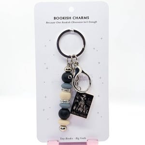 May include: A keychain with a beaded charm and a miniature book. The charm features black, gray, and cream-colored beads, along with silver accents. The tiny book reads "The Ritual" by Shantel Tessier. The card reads "Bookish Charms" and "Tiny Books - Big Feels."