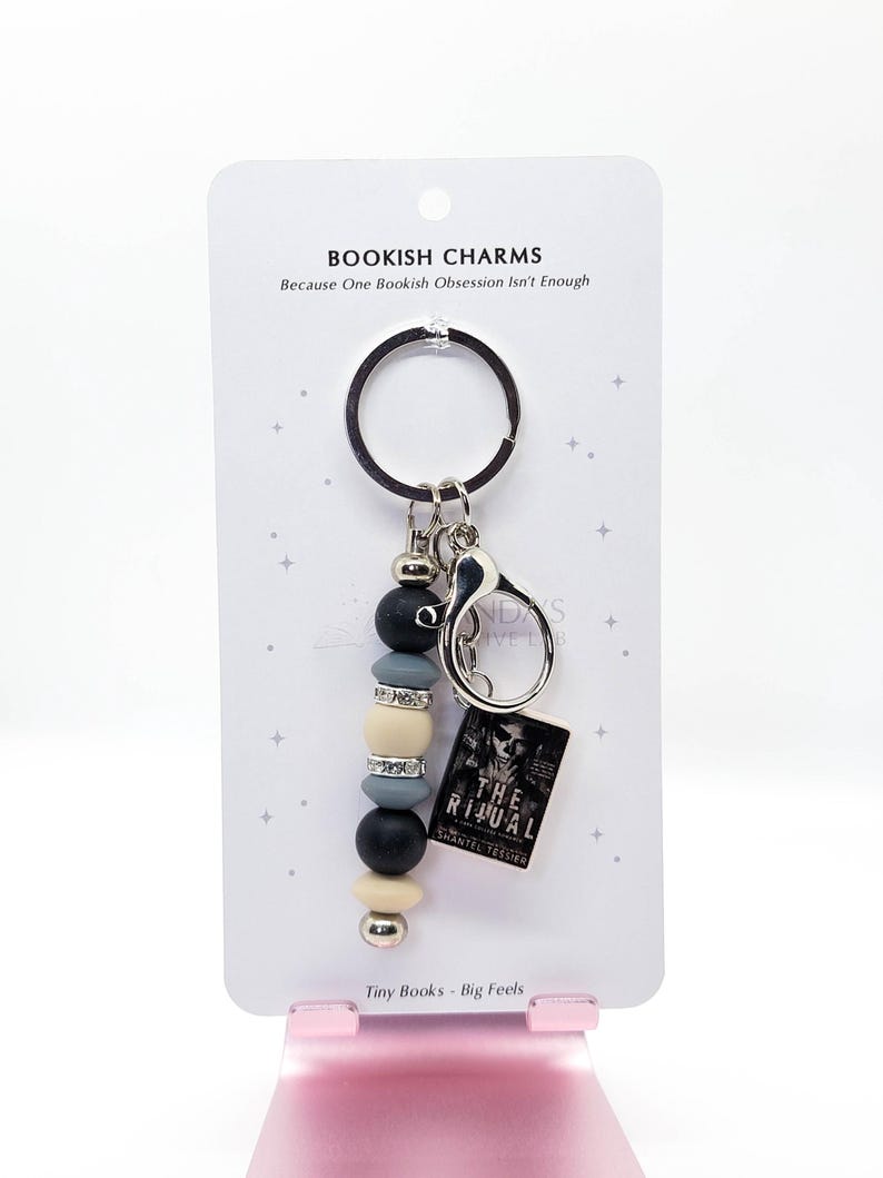 May include: A keychain with a beaded charm and a miniature book. The charm features black, gray, and cream-colored beads, along with silver accents. The tiny book reads "The Ritual" by Shantel Tessier. The card reads "Bookish Charms" and "Tiny Books - Big Feels."