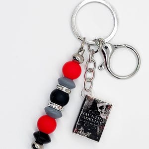 May include: A keychain with a silver ring and clasp. The keychain features a beaded charm with red, gray, black, and silver beads. A small book charm with the title "Haunting Adeline" is also attached.