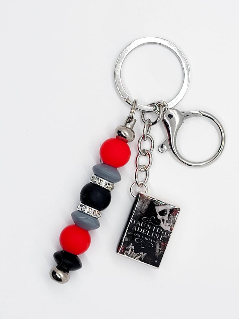 May include: A keychain with a silver ring and clasp. The keychain features a beaded charm with red, gray, black, and silver beads. A small book charm with the title "Haunting Adeline" is also attached.