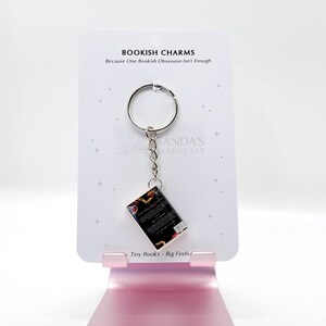 May include: A keychain featuring a miniature book charm. The charm is attached to a silver keyring and a chain. The card behind the charm reads "BOOKISH CHARMS" and "Tiny Books - Big Feels."