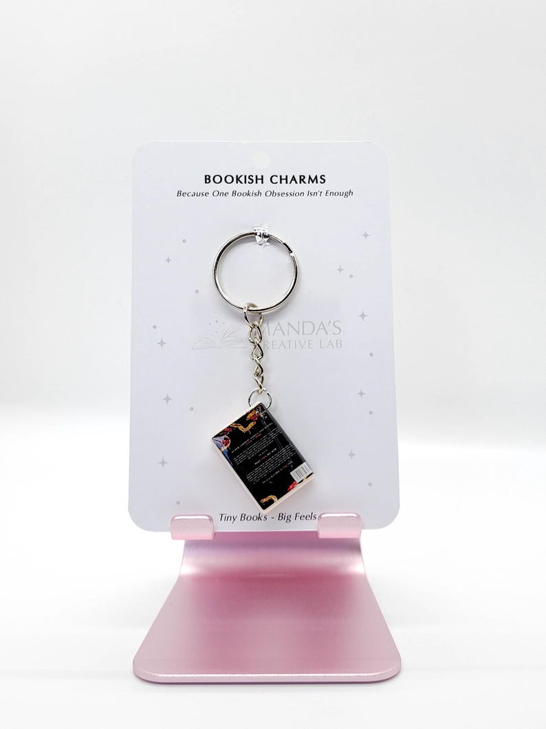 May include: A keychain featuring a miniature book charm. The charm is attached to a silver keyring and a chain. The card behind the charm reads "BOOKISH CHARMS" and "Tiny Books - Big Feels."