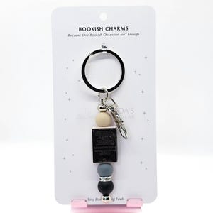May include: A keychain with a black ring and a silver clasp. The keychain features a cream-colored bead, a small black book, a gray bead, and a black bead. The card behind the keychain reads "BOOKISH CHARMS Because One Bookish Obsession Isn't Enough."