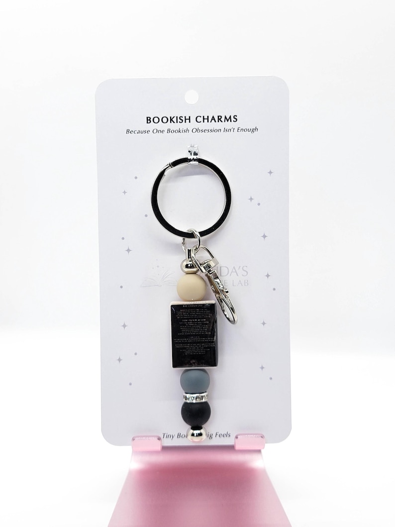 May include: A keychain with a black ring and a silver clasp. The keychain features a cream-colored bead, a small black book, a gray bead, and a black bead. The card behind the keychain reads "BOOKISH CHARMS Because One Bookish Obsession Isn't Enough."