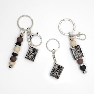 May include: Keychains with a small book charm that reads "The Sinner". The keychains have silver-colored rings and chains. Some keychains have decorative beads in shades of brown, black, and cream.