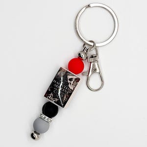 May include: A silver keychain with a lobster clasp and a key ring. The keychain features a small book charm with the text "Haunting Adeline" and a skull graphic. It also includes red, black, gray, and silver beads.