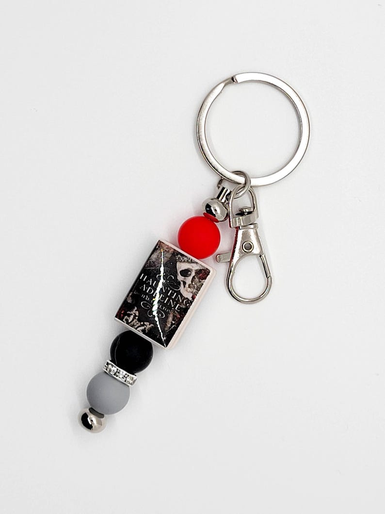 May include: A silver keychain with a lobster clasp and a key ring. The keychain features a small book charm with the text "Haunting Adeline" and a skull graphic. It also includes red, black, gray, and silver beads.