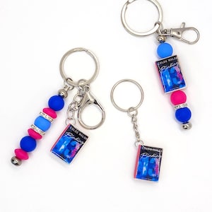 May include: Keychains with book charms and colorful beads. The charms feature the book cover of "Binding" by Chloe Walsh. The keychains have silver-colored metal rings and clasps, with pink, blue, and silver beads.