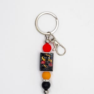 May include: A keychain with a silver ring and clasp. The keychain features a rectangular charm with a snake and rose design, along with red, orange, black, and silver beads. The charm has the text "ORY VERSES".