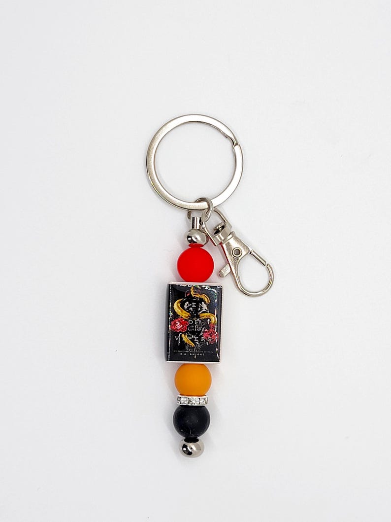 May include: A keychain with a silver ring and clasp. The keychain features a rectangular charm with a snake and rose design, along with red, orange, black, and silver beads. The charm has the text "ORY VERSES".